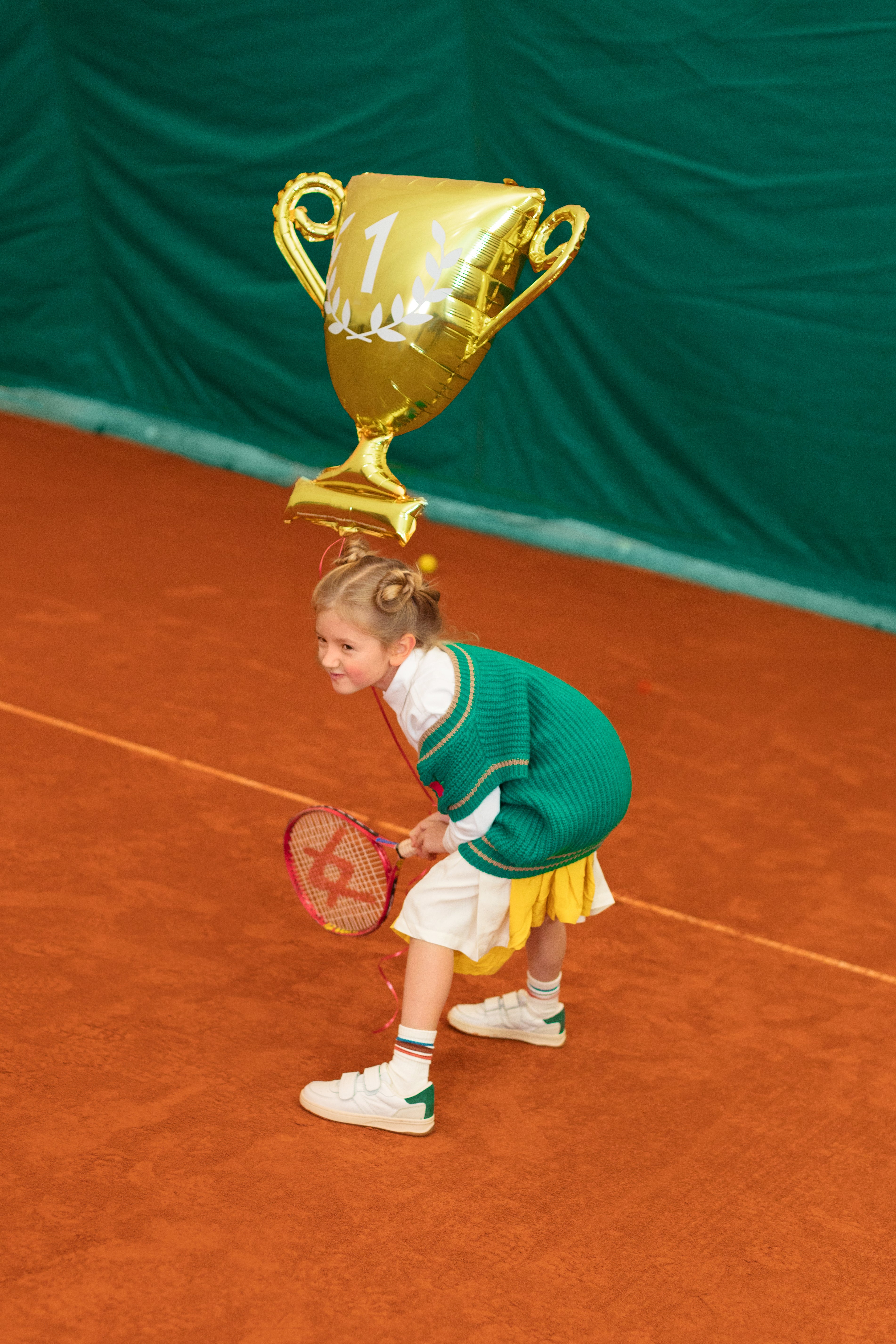 Child on a tennis court with a large gold trophy above their head