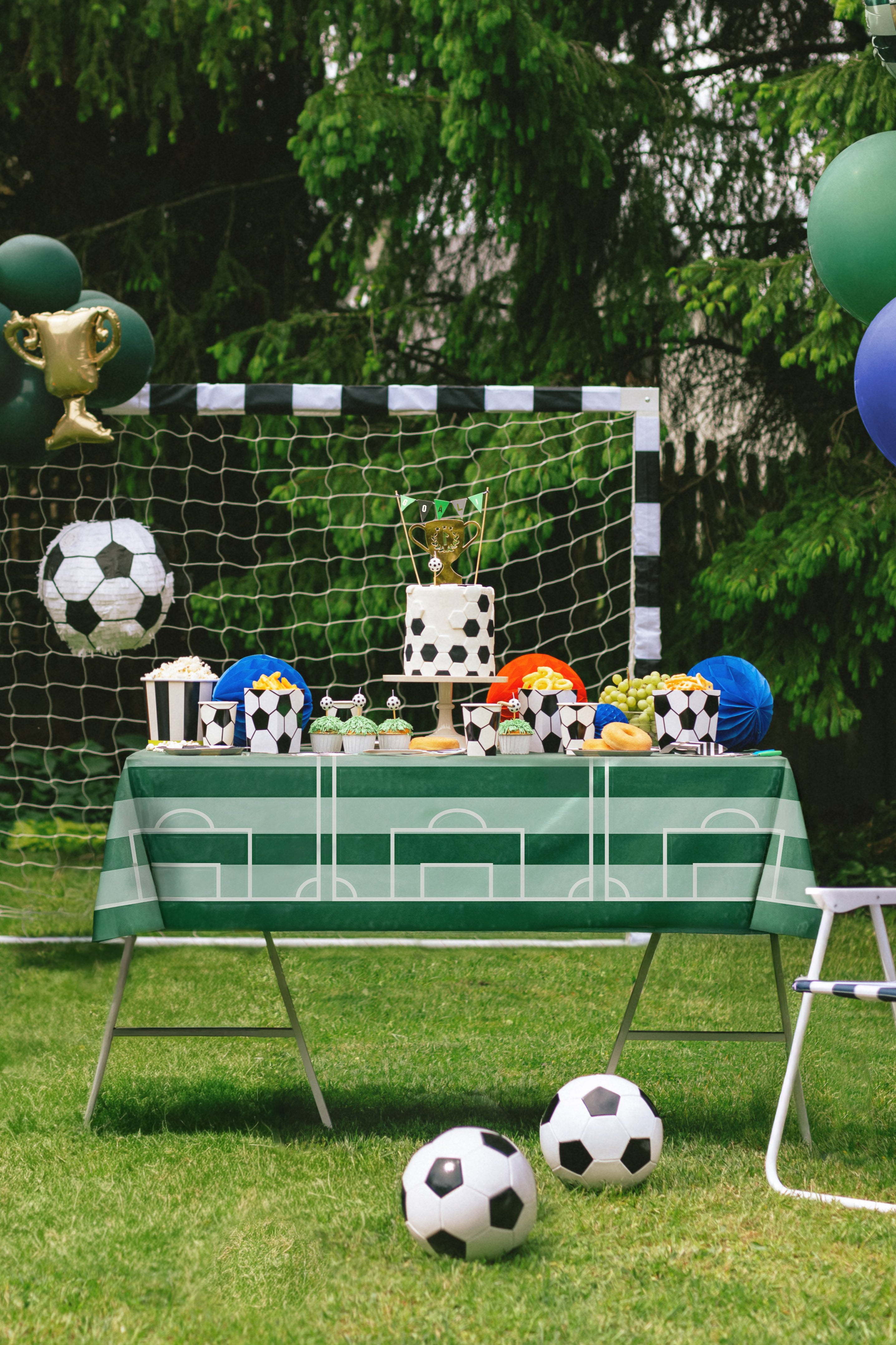 Children's party setup with soccer-themed decorations, including a table with small cakes, balloons, and soccer balls on a grassy outdoor area.