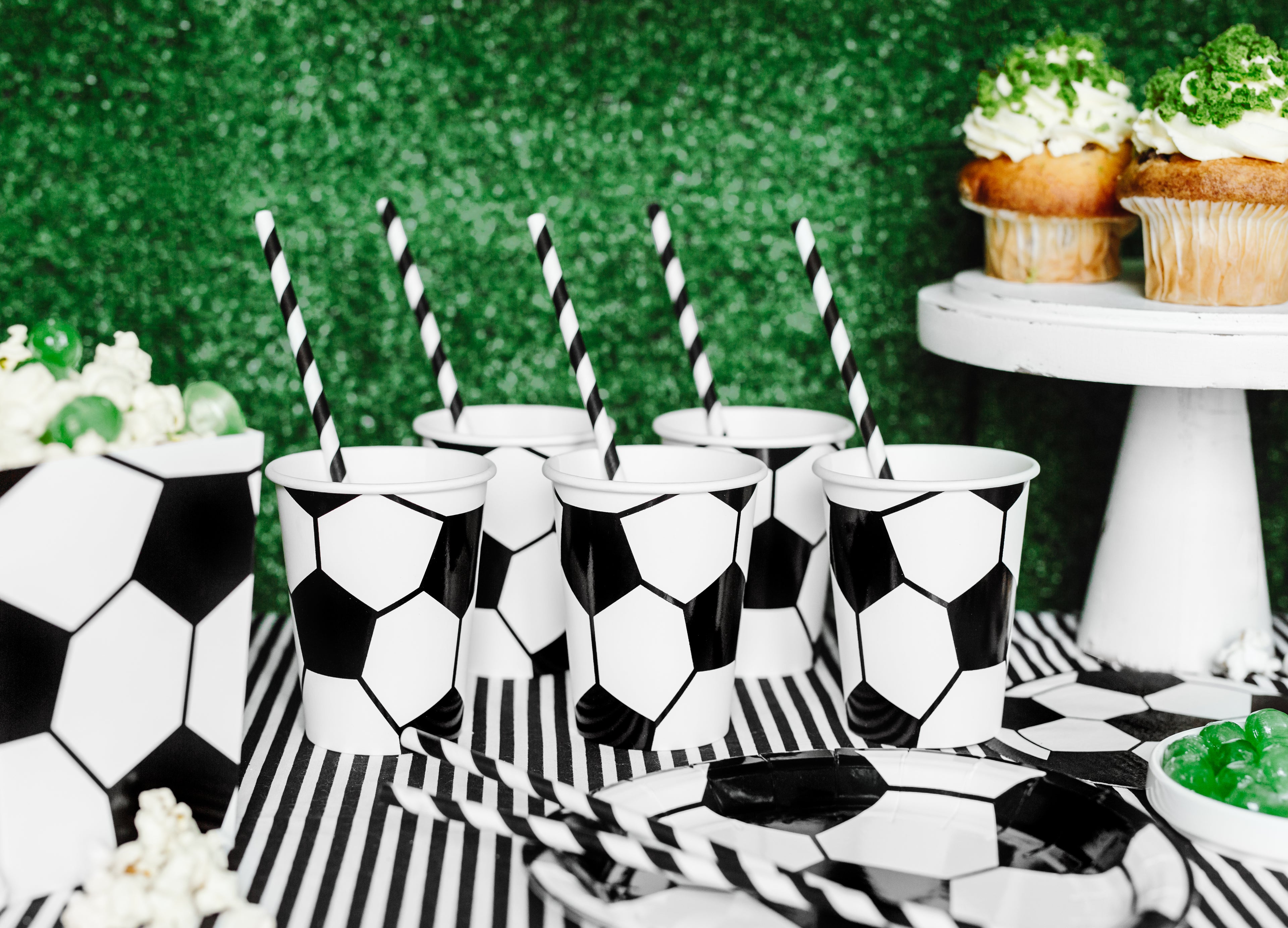 Soccer-themed party with black and white cups, striped straws, and greenery in the background.