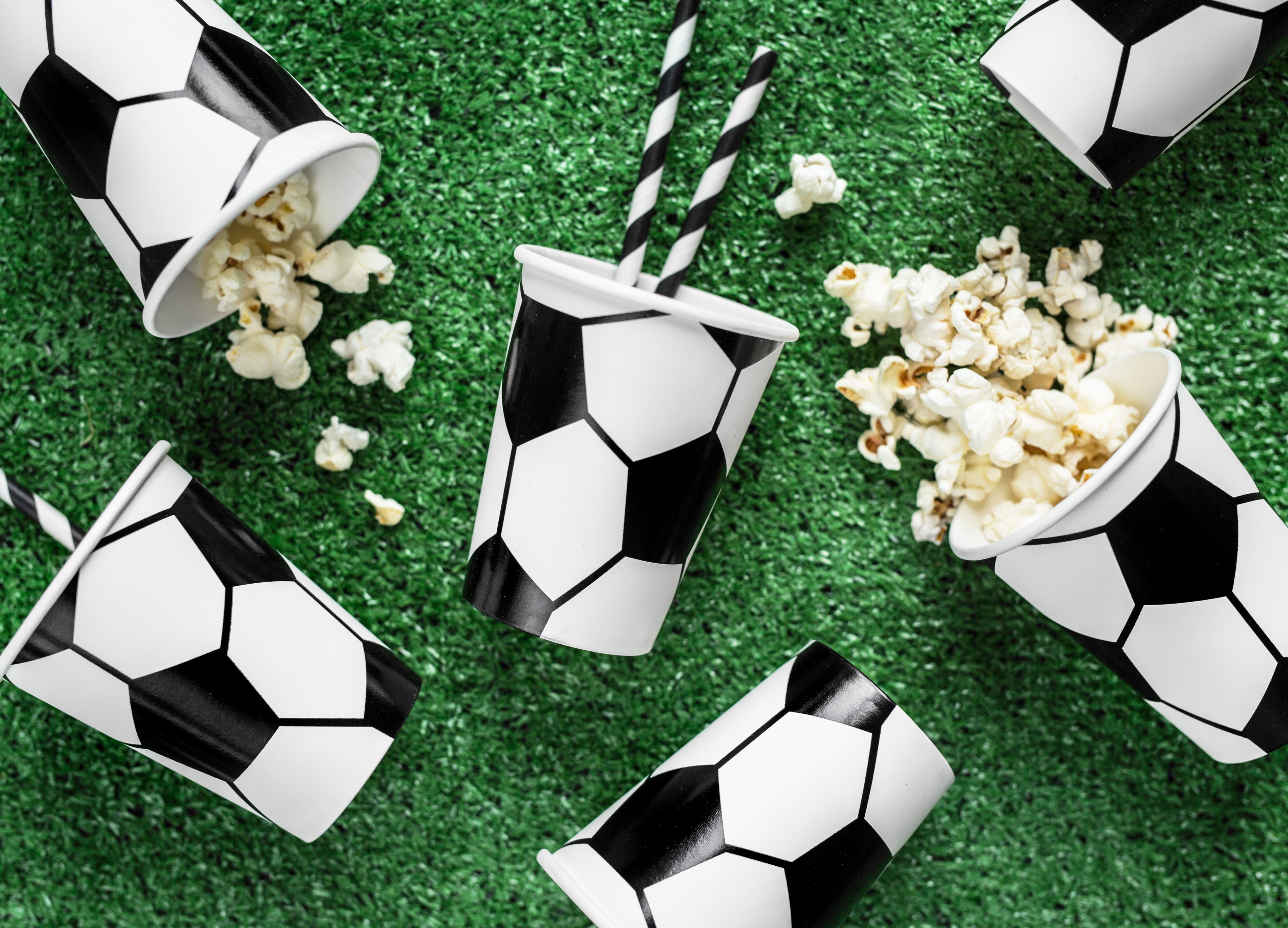 Soccer-themed popcorn cups with black and white striped straws on a green surface.