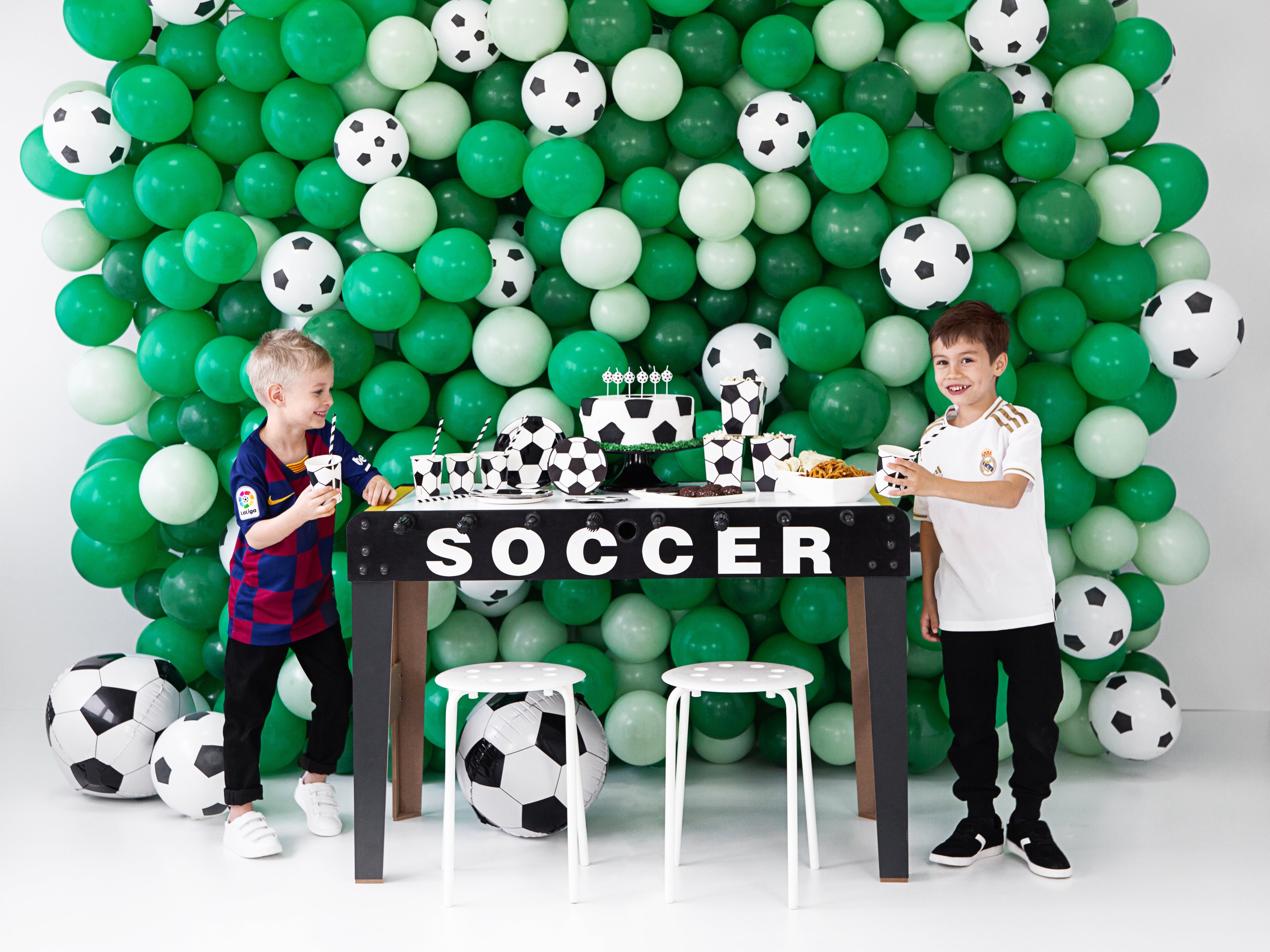 Two children standing next to a table with soccer-themed decorations in front of a green and white balloon display.
