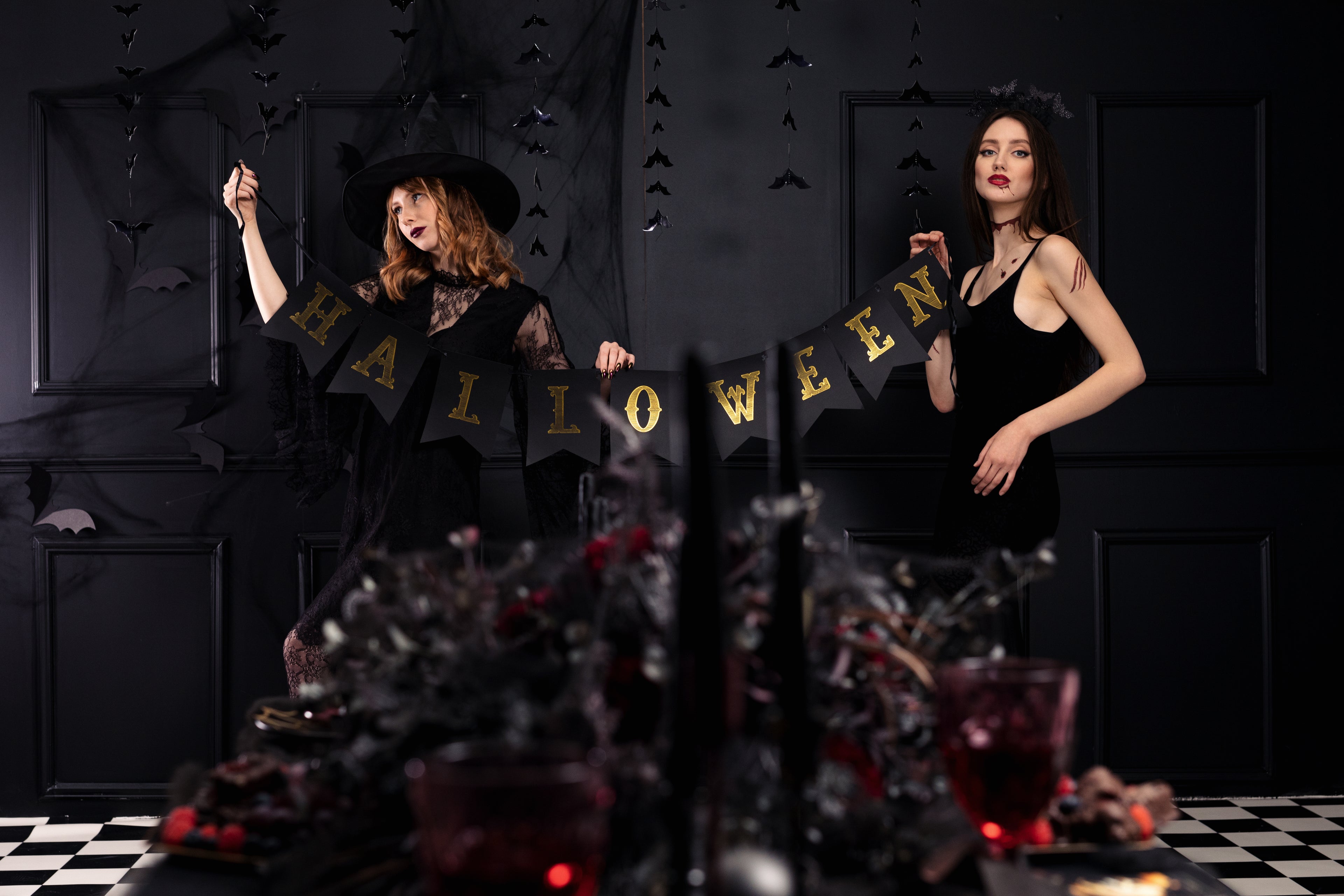 Two women in Halloween costumes standing behind a table with a 'Halloween' banner in a darkly decorated room.