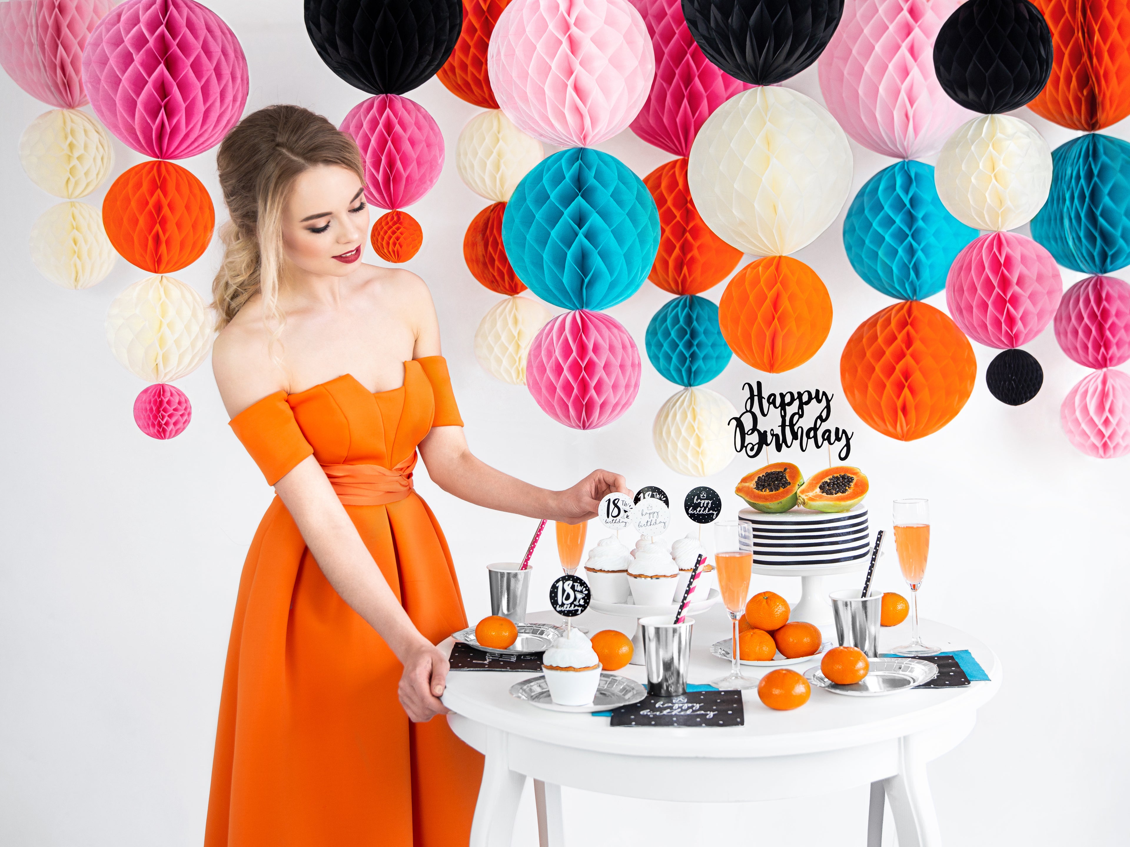 Woman in an orange dress standing next to a table with a birthday cake and decorations, surrounded by colorful paper balls.